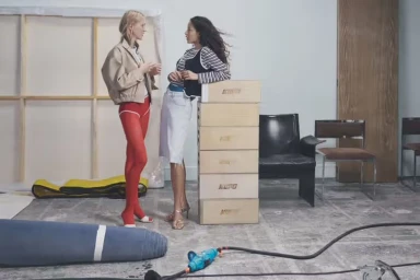 Two models standing and talking inside a studio with stacked wooden boxes and chairs in the background