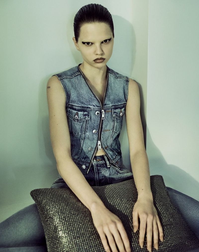Portrait of a young adult female wearing a denim vest and jeans, seated indoors with a pillow, looking at the camera with a serious expression.