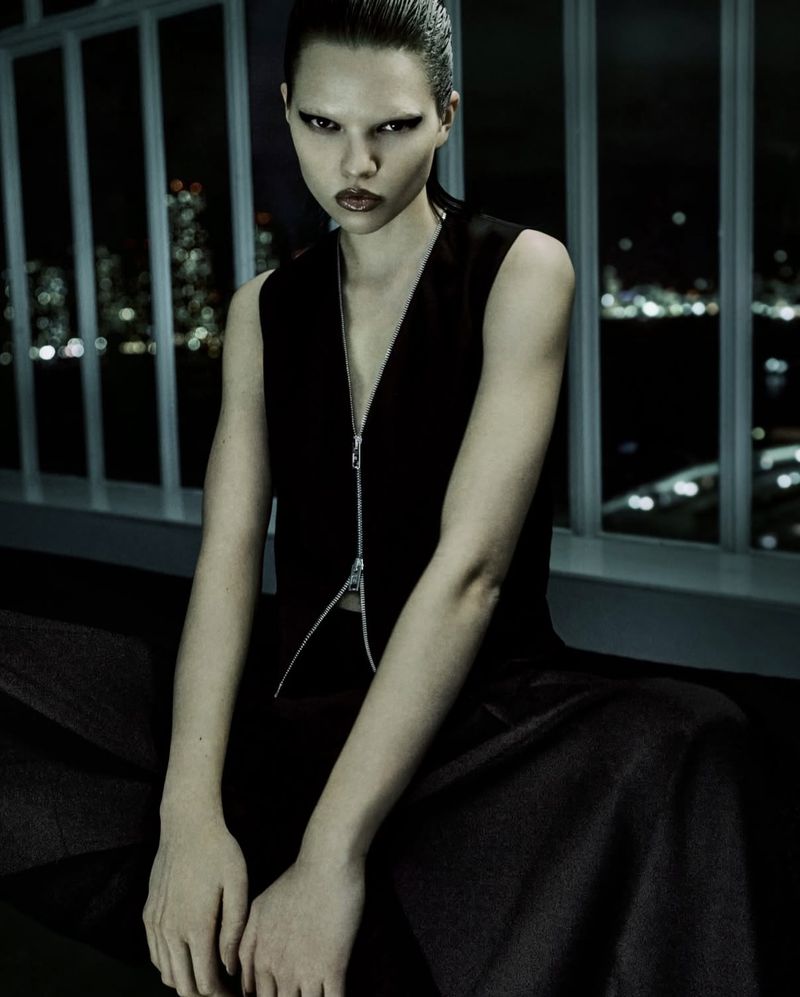 Dark fashion portrait of a woman with slicked-back hair wearing a black vest against a blurred cityscape background at night.