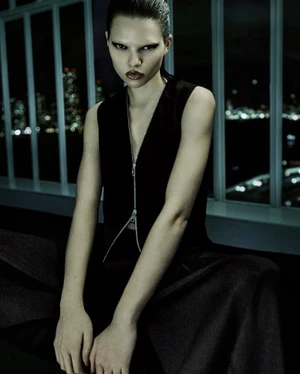 Model wearing a sleeveless black zippered vest and dark trousers against a window with blurred city lights at night.