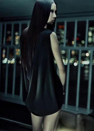 Model with long dark hair wearing a black sleeveless dress posing indoors against a railing with city lights in the background.