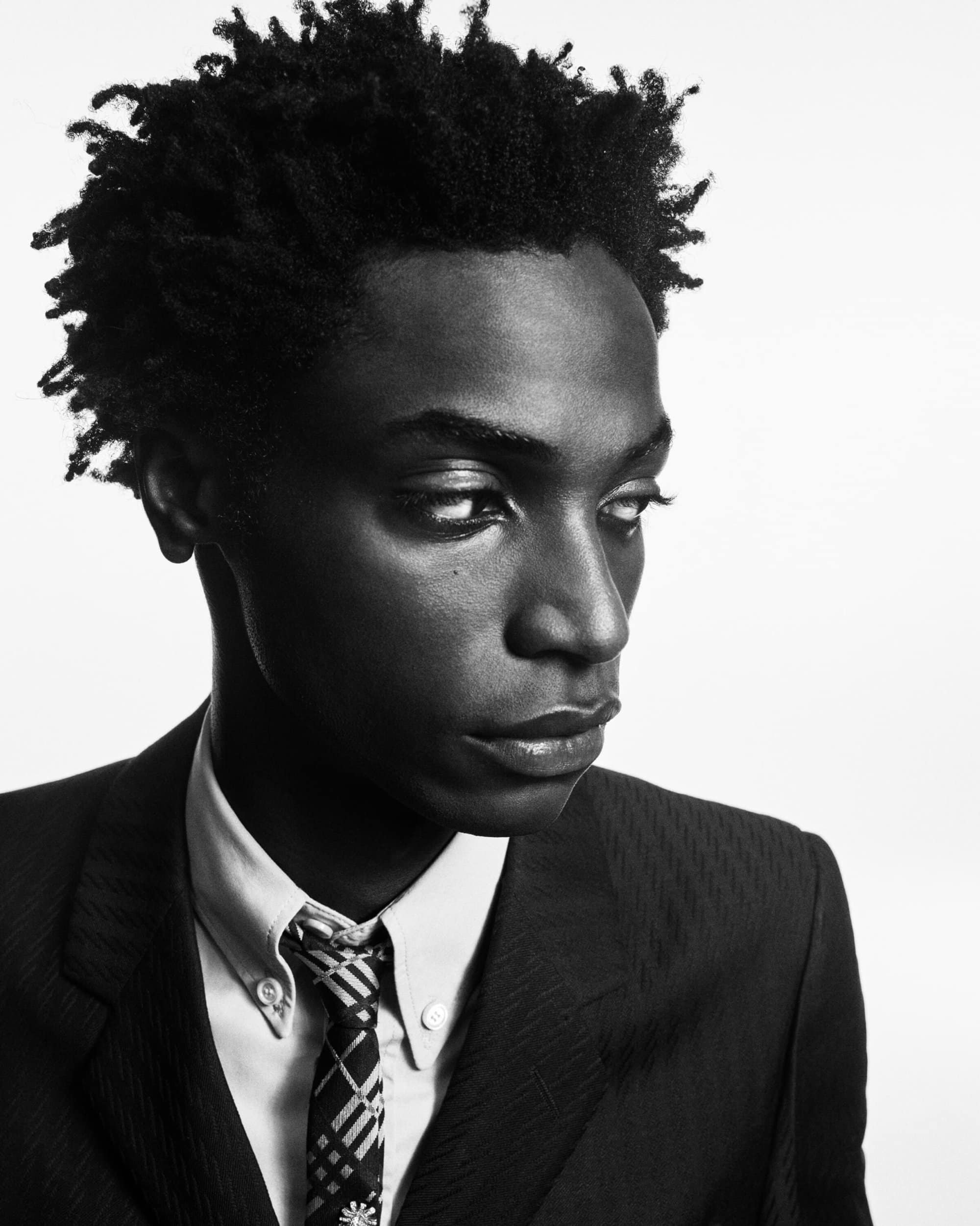 Black and white portrait of a Black male in a suit and tie looking off to the side against a neutral background in eye-level framing.