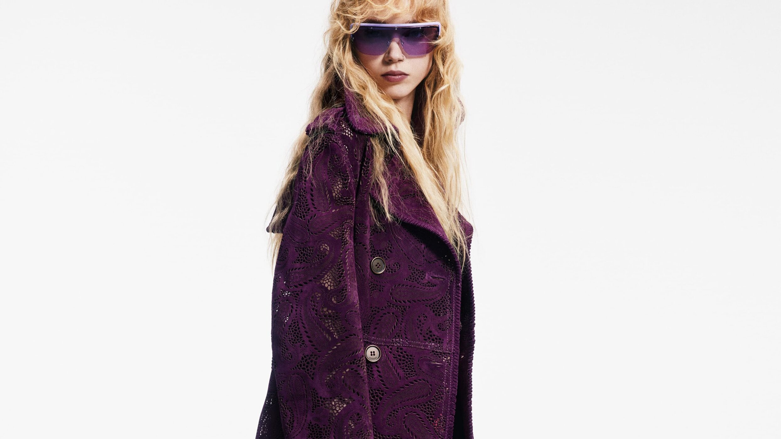 Adult female with long wavy hair poses wearing sunglasses and a patterned purple trench coat in a bright, high-key photo studio.