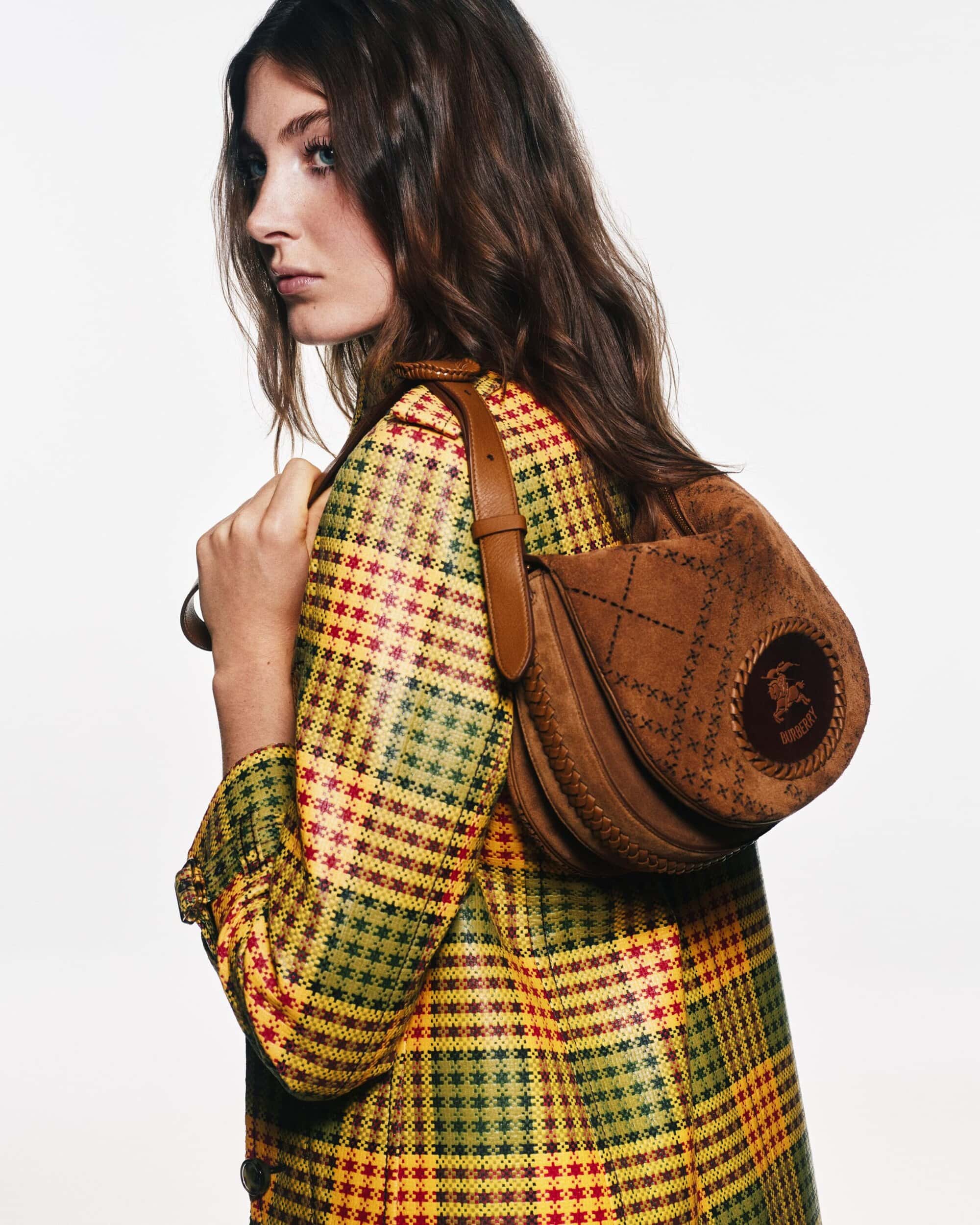 A woman wearing a yellow and red plaid coat carrying a brown suede shoulder bag against a white background.
