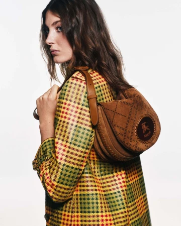 A woman wearing a yellow and red plaid coat carrying a brown suede shoulder bag against a white background.