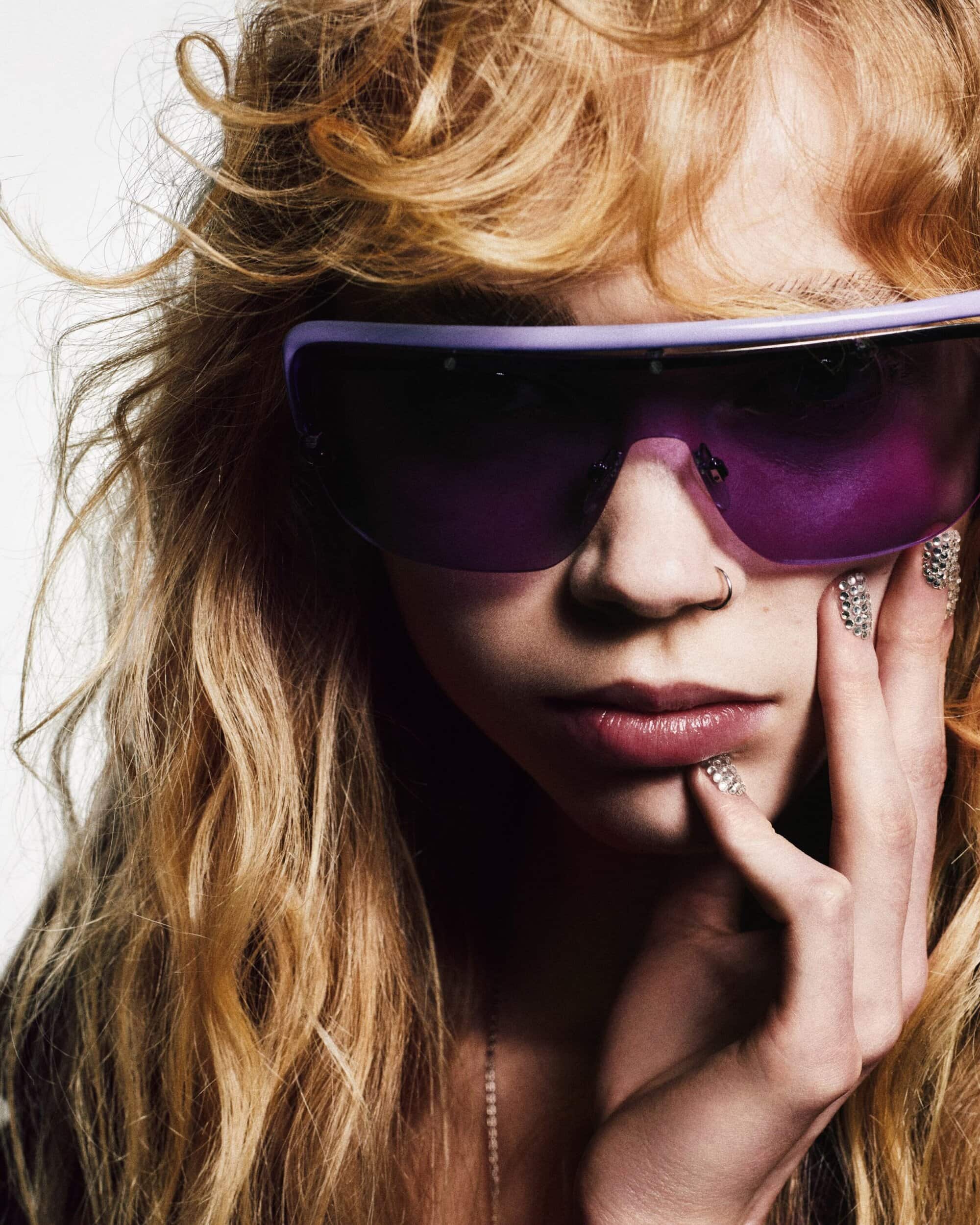 Close-up portrait of a young woman wearing purple sunglasses and crystal-embellished nail art against a white background.