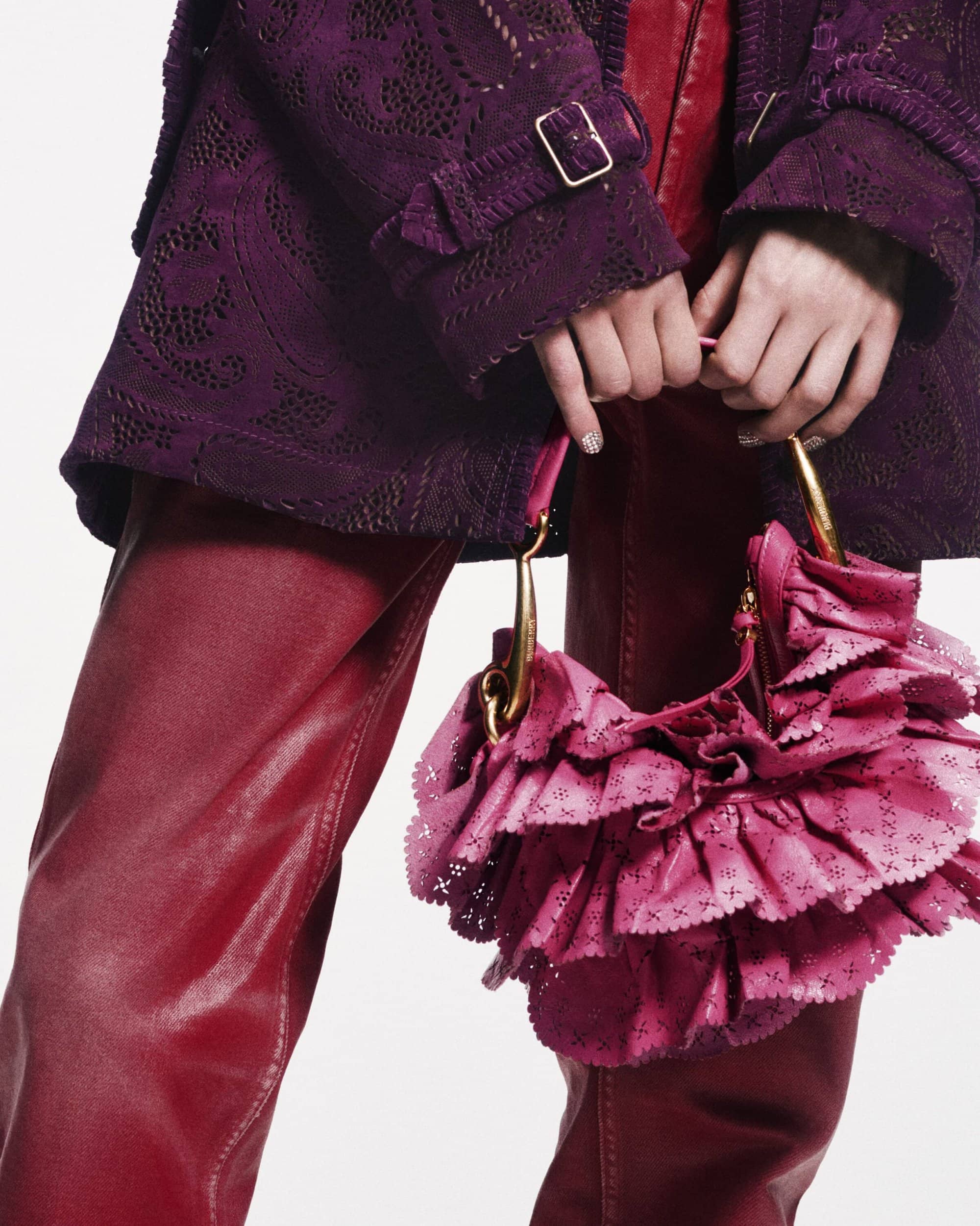 Close-up of a fashion model's hands holding a pink ruffled handbag, wearing a purple lace jacket and red leather pants in a studio.