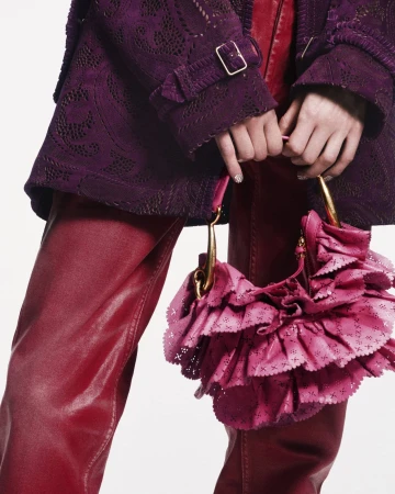 Close-up of a fashion model's hands holding a pink ruffled handbag, wearing a purple lace jacket and red leather pants in a studio.