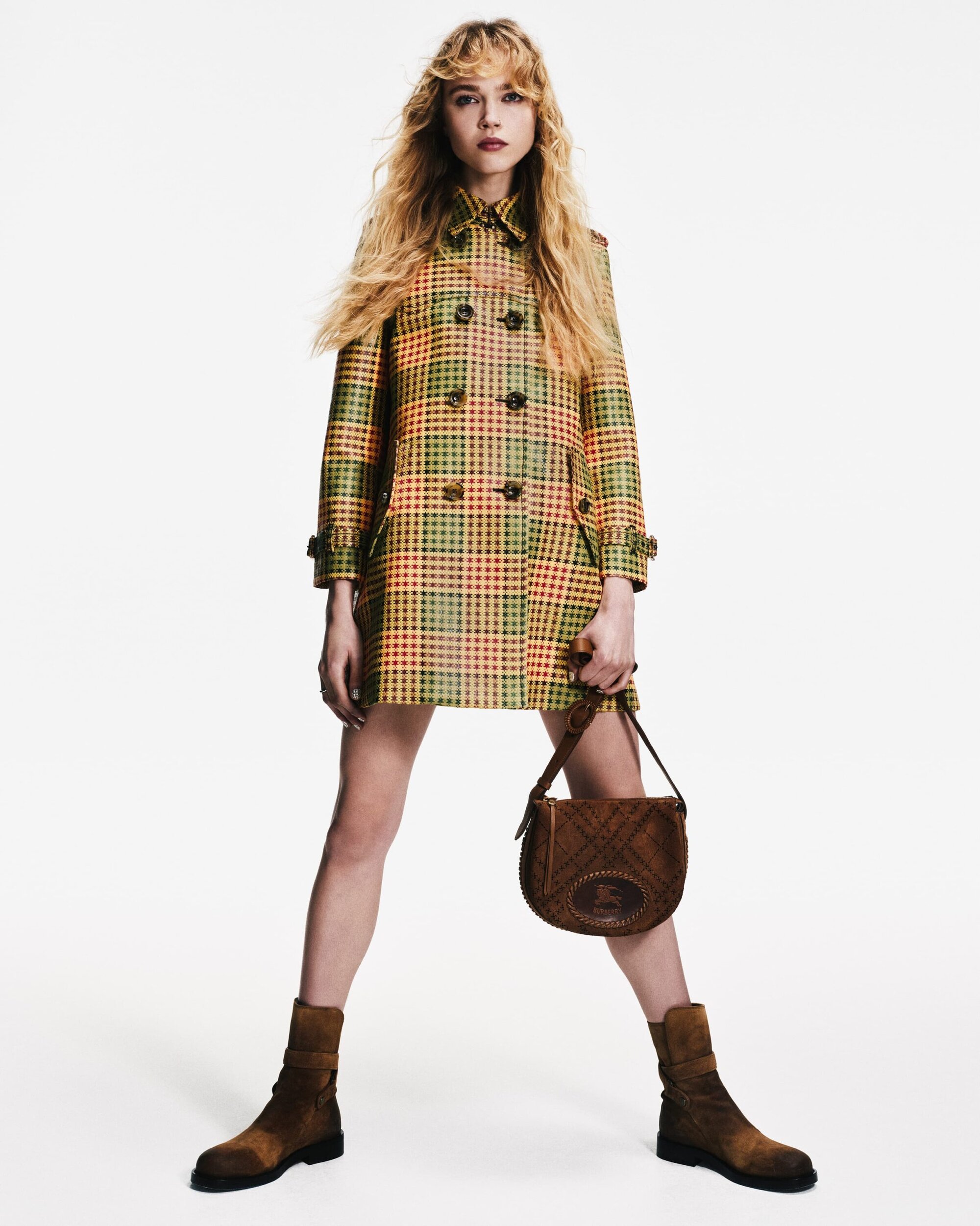 A woman in a plaid coat, brown boots, and handbag stands against a white background in a fashion-style full shot.