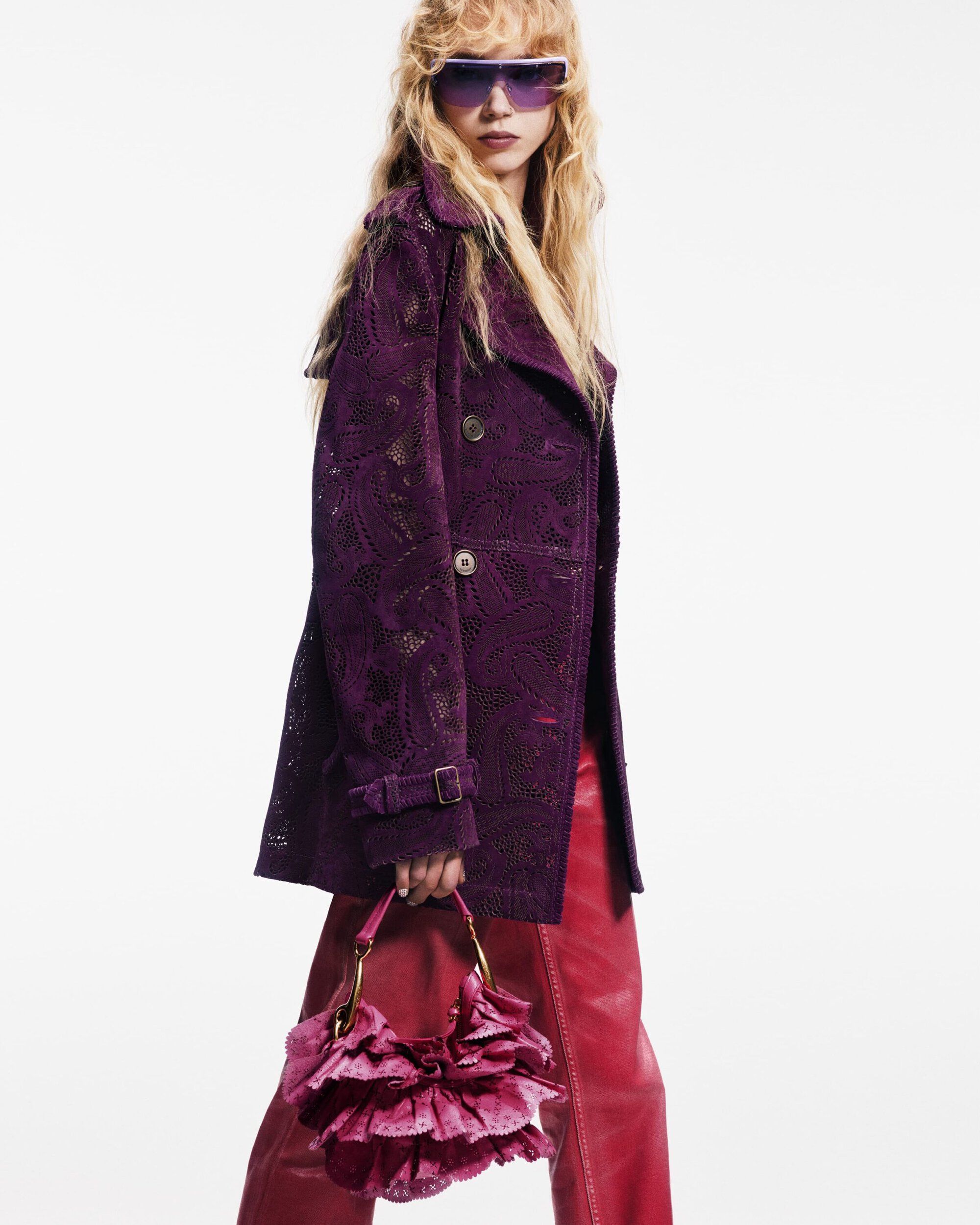 A blonde woman poses in a plum jacket and red leather pants, holding a ruffled purse against a white background in a fashion shot.
