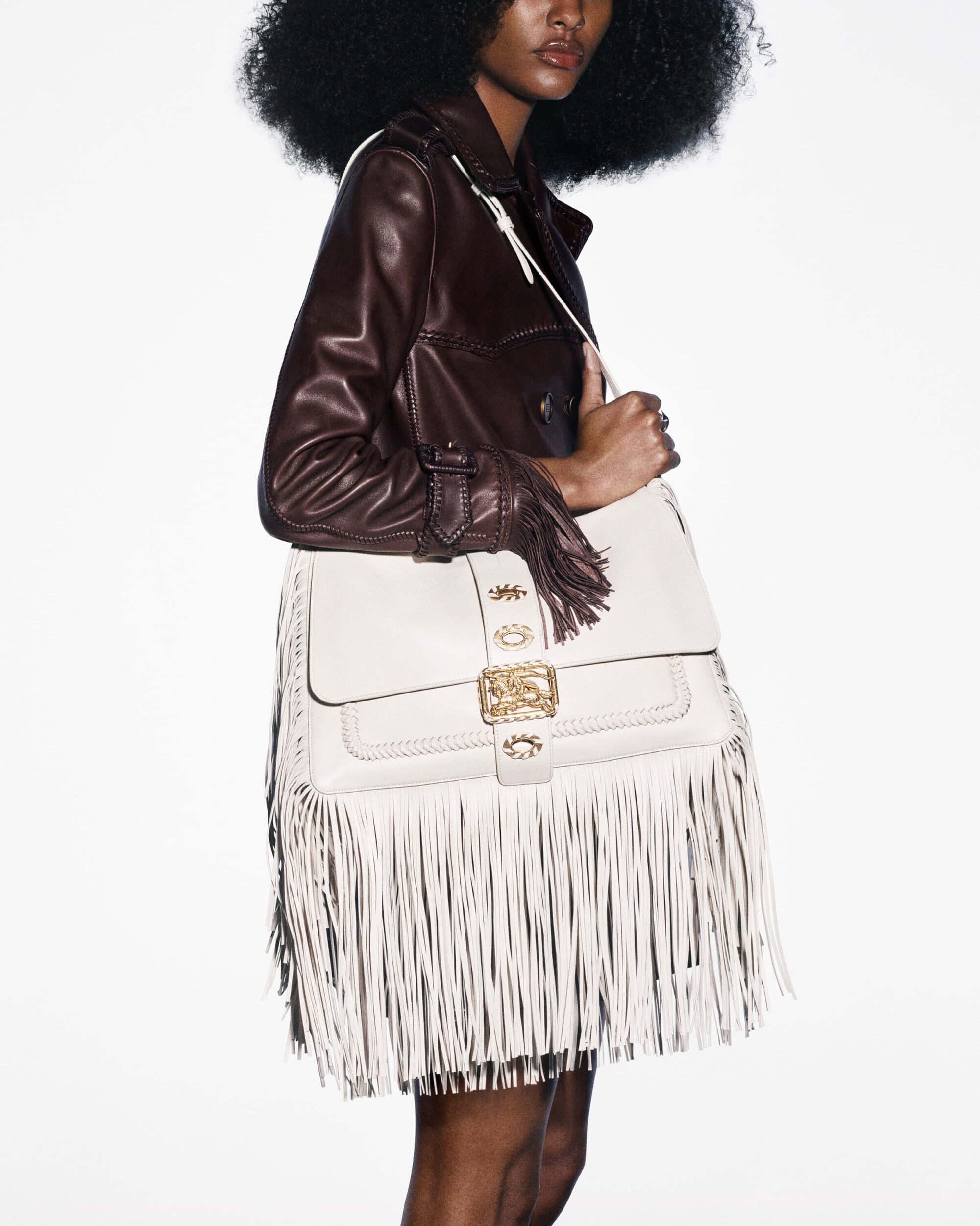 Dark-skinned model showcases a white leather fringe purse and a brown leather jacket on a white background in a fashion editorial shot.