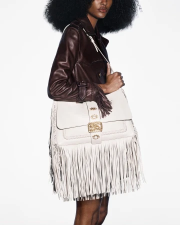 Dark-skinned model showcases a white leather fringe purse and a brown leather jacket on a white background in a fashion editorial shot.