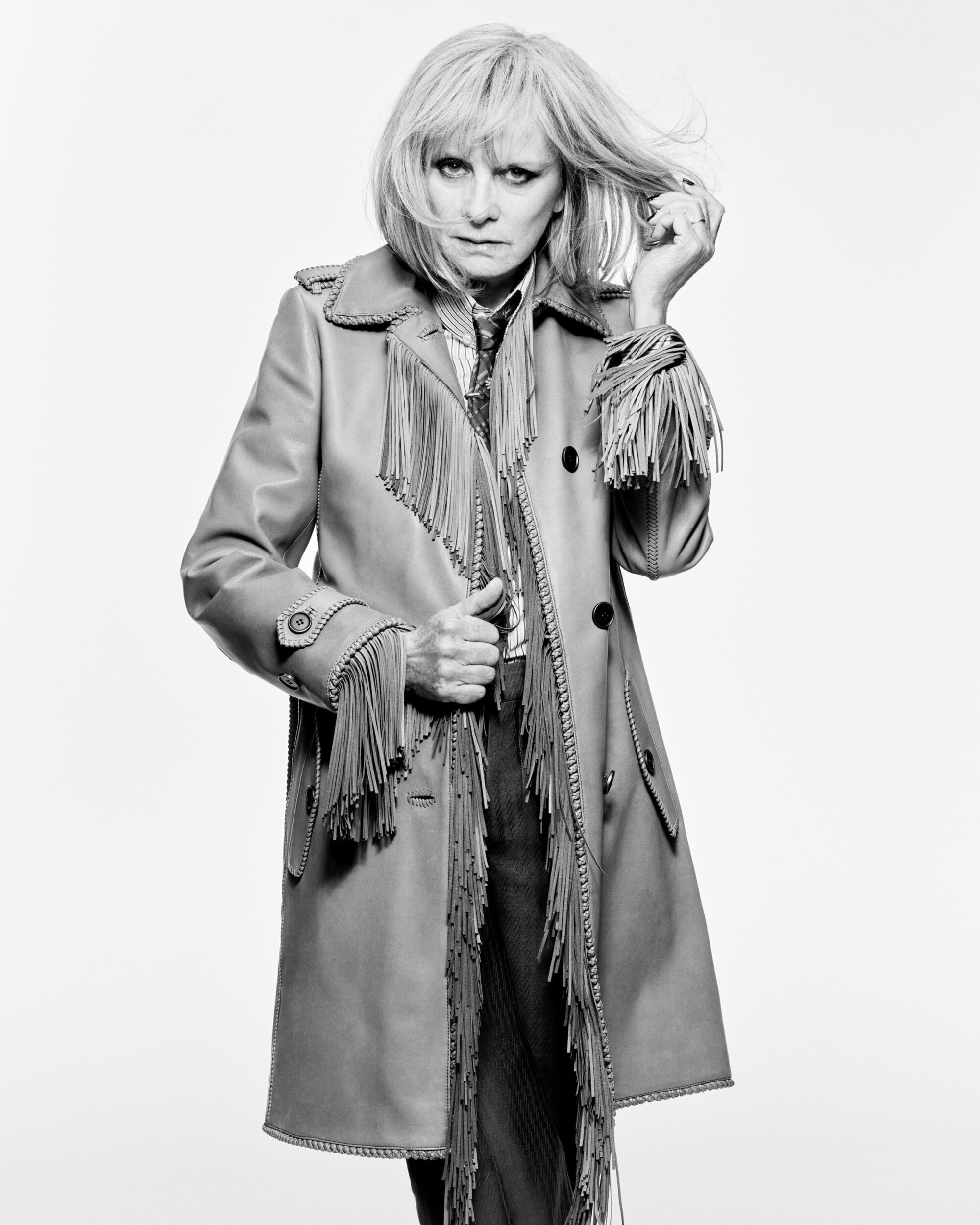Black and white studio shot of a woman with blonde hair wearing a fringe leather coat. She is holding her hair and looking at the camera.