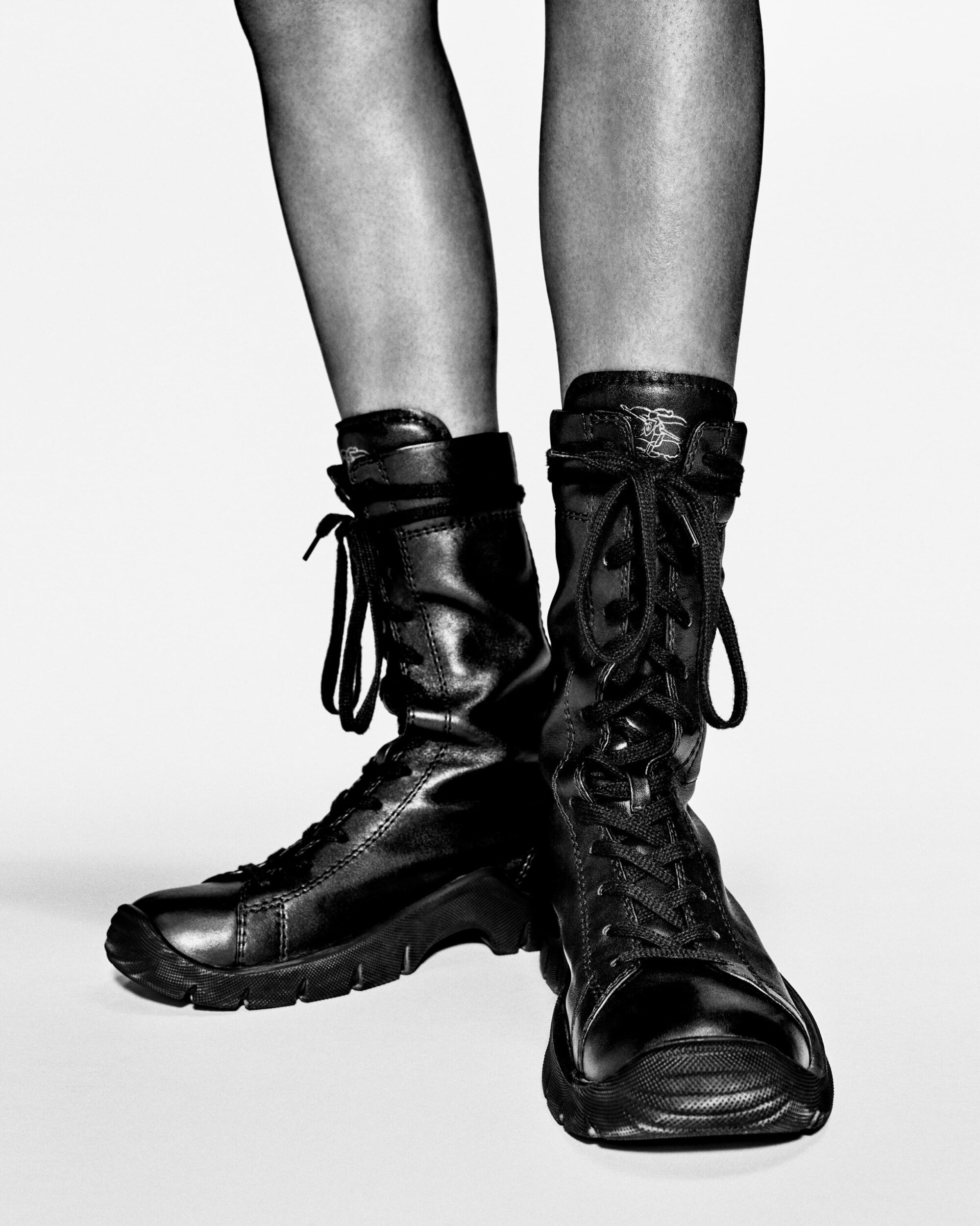 Close-up of person wearing black leather combat boots and stockings on white background in studio lighting, fashion footwear product shot.