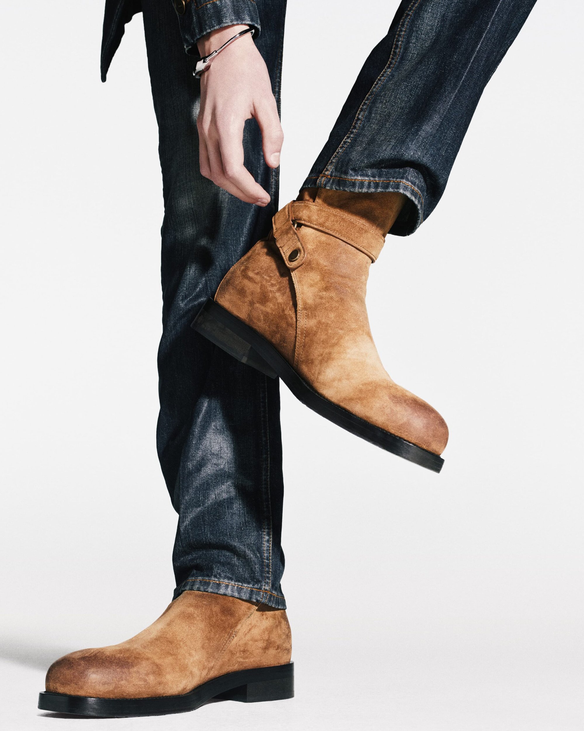 Closeup of a man's lower legs, showing the brown suede boots he is wearing with dark blue jeans in a bright studio setting.