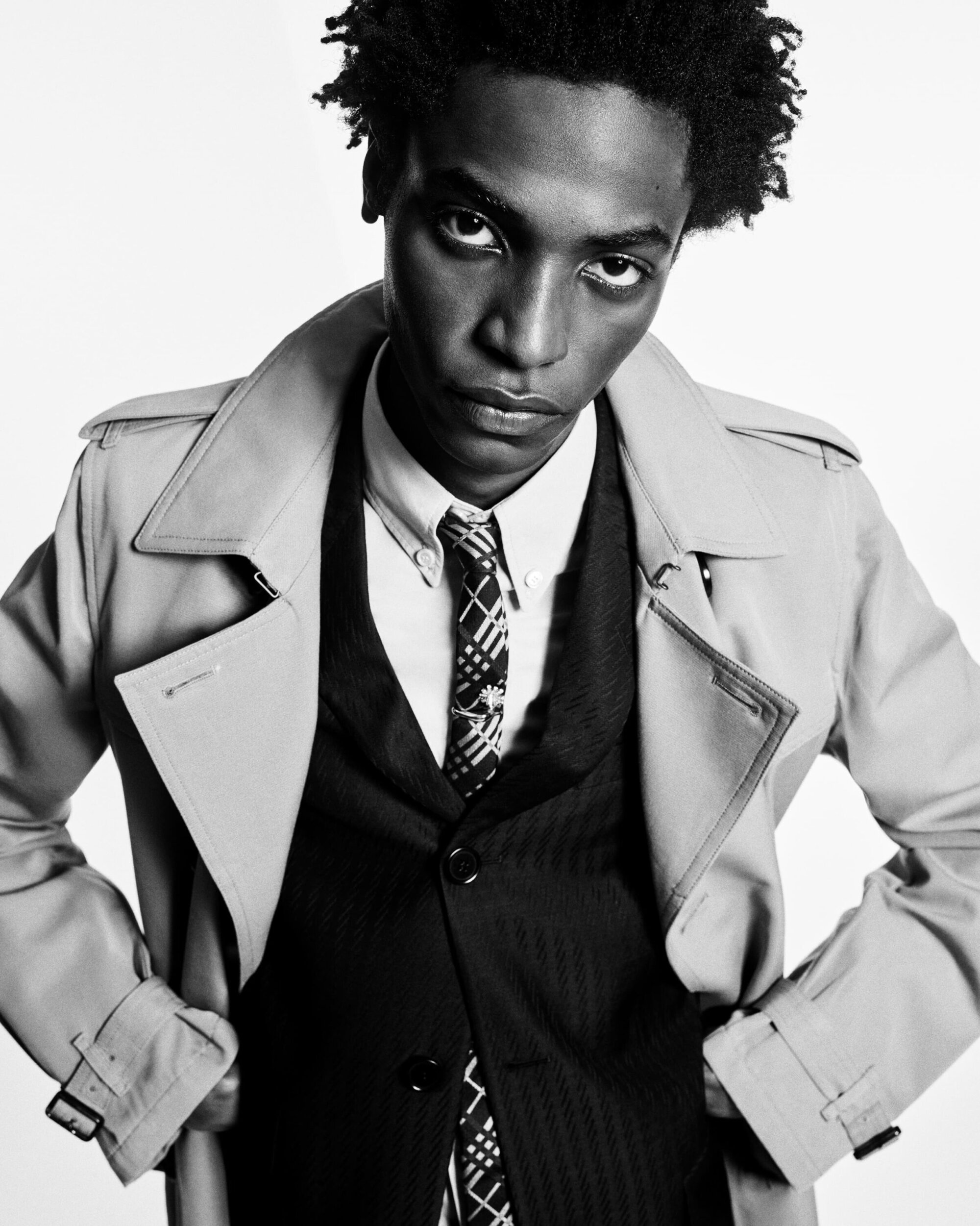 Black-and-white portrait of a Black male model wearing a tan trench coat, patterned tie, white collared shirt, and dark vest against white.