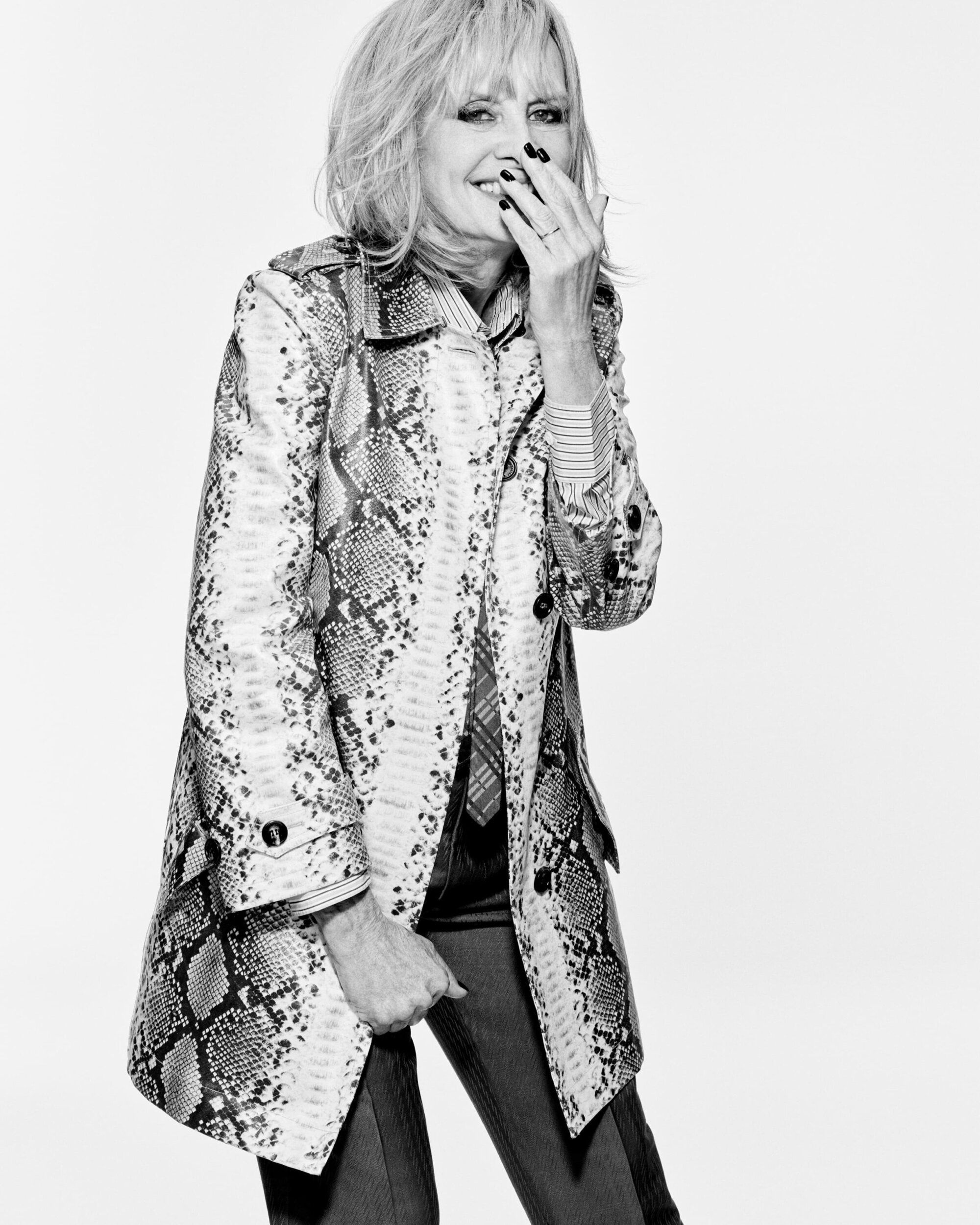 A black-and-white portrait of a mature woman with blonde hair in a snakeskin coat, smiling with hand near face against a light backdrop.