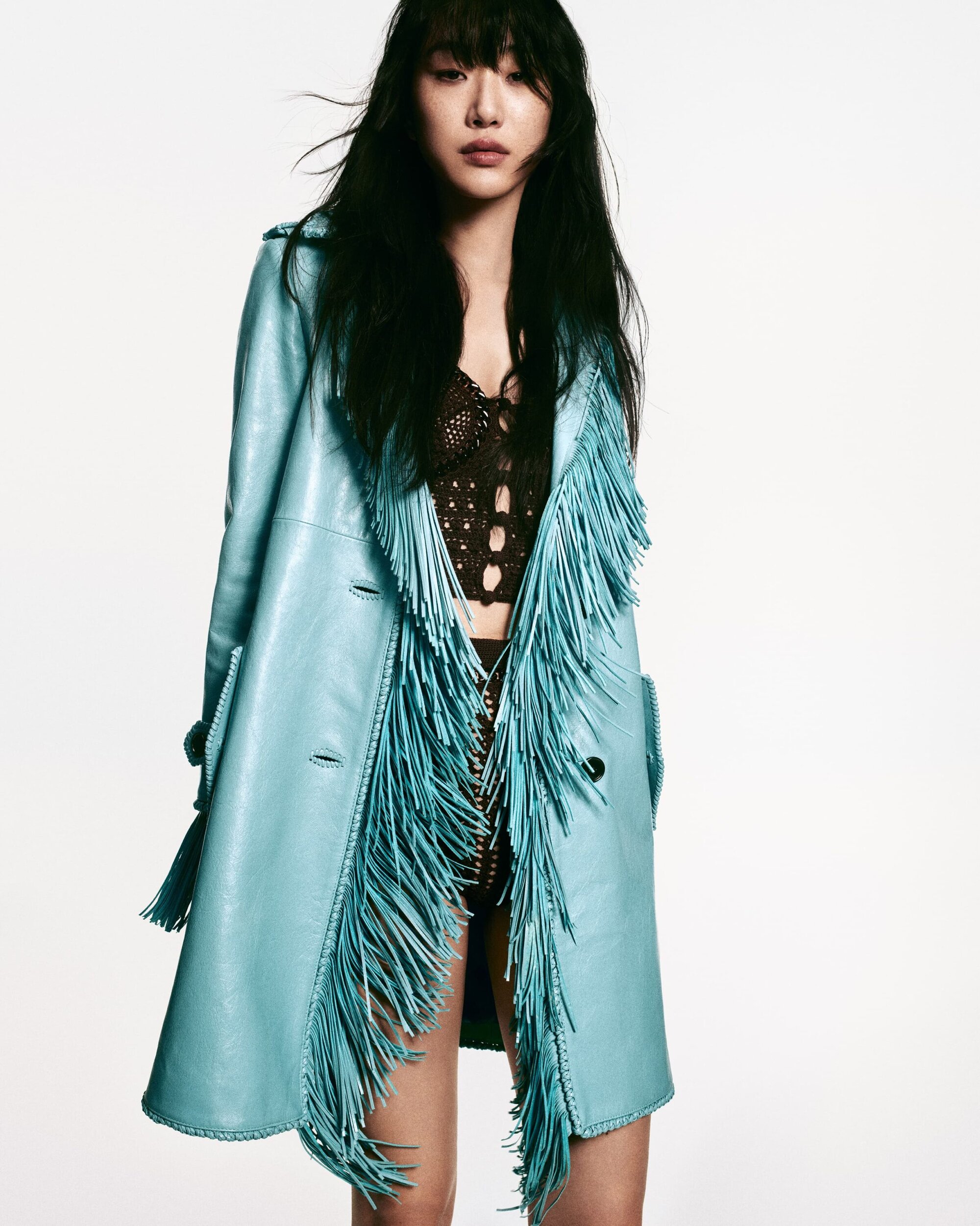 Fashion shot of a woman wearing a turquoise fringe coat and black crochet top, against a clean white backdrop in a photo studio.