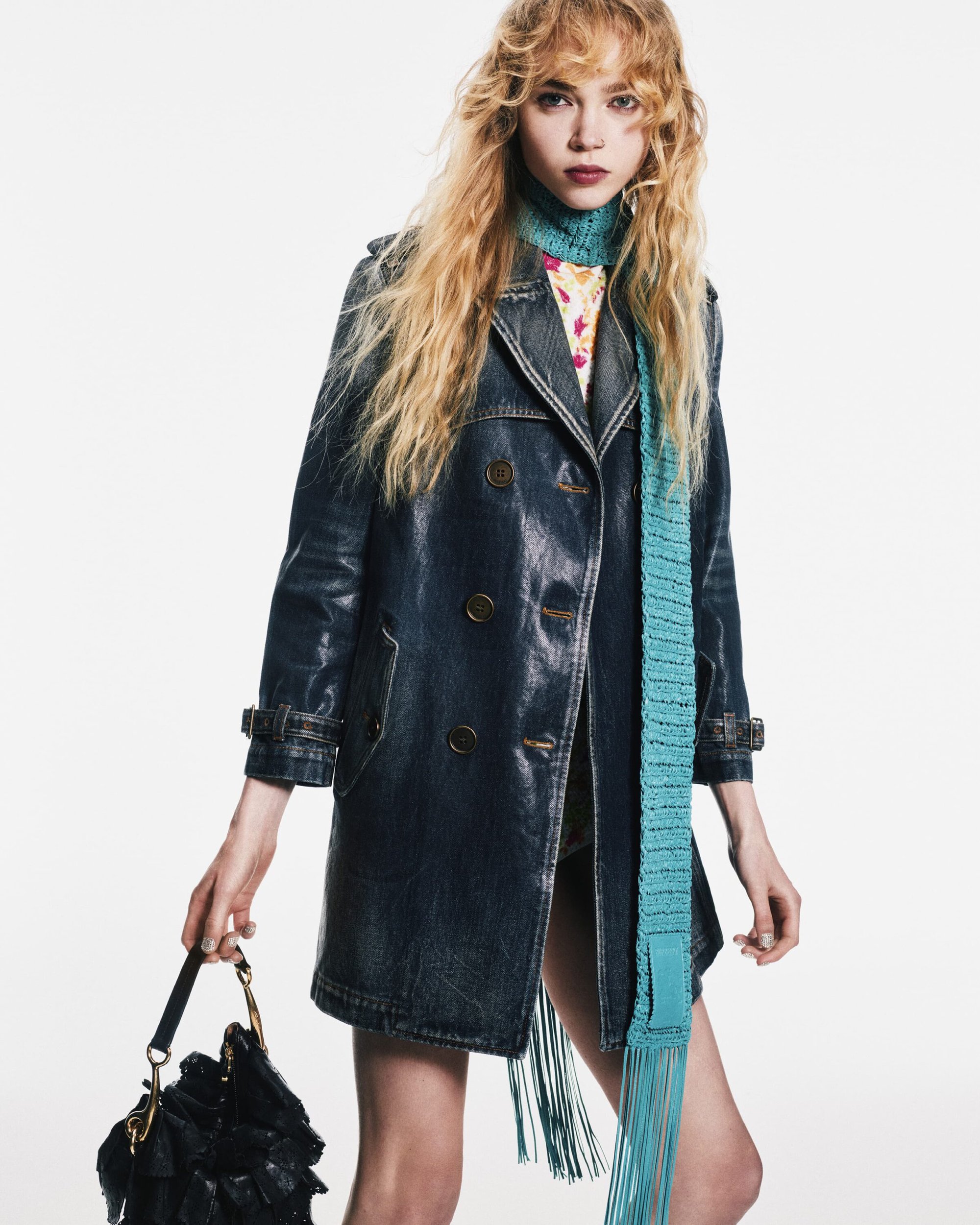 Fashion shot of woman in blue coat and teal scarf holding a black bag against white background, studio lighting. Editorial, full body.