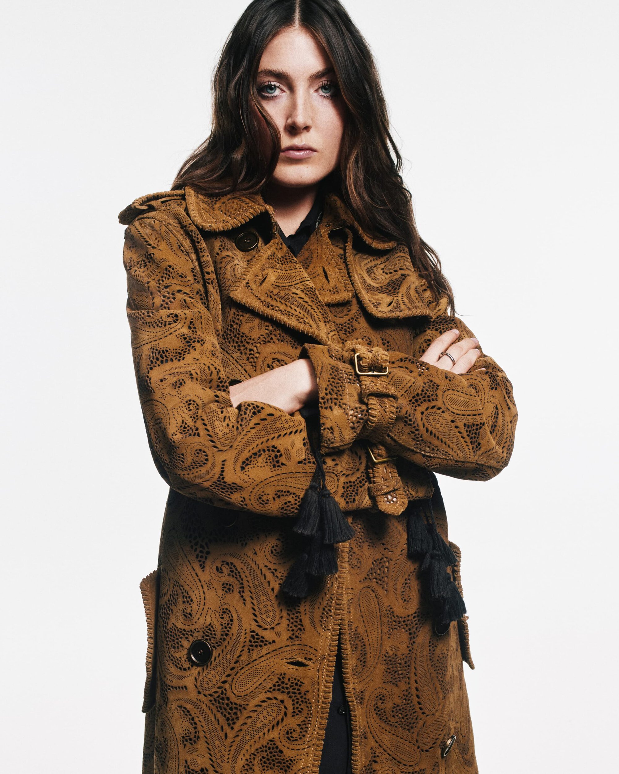 Brunette woman in patterned brown trench coat with arms crossed in a fashion studio shot, styled hair, eye-level angle, on white backdrop.