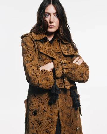 Brunette woman in patterned brown trench coat with arms crossed in a fashion studio shot, styled hair, eye-level angle, on white backdrop.