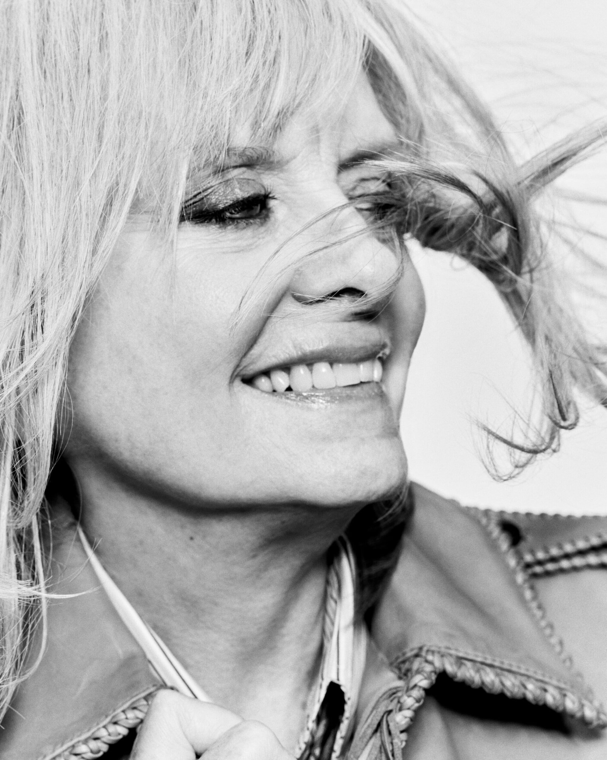 Close-up black and white portrait of a blonde woman with windblown hair smiling and wearing a jacket against a bright background.