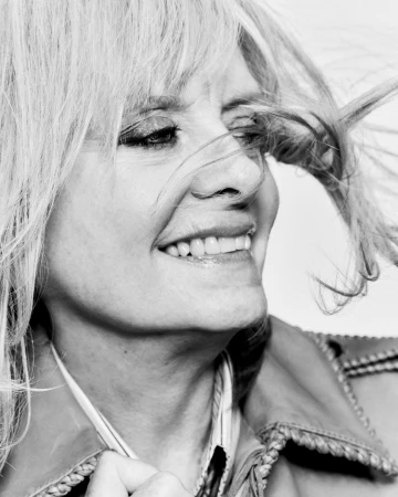 Close-up black and white portrait of a blonde woman with windblown hair smiling and wearing a jacket against a bright background.