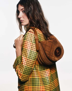 A woman wearing a yellow and red plaid coat carrying a brown suede shoulder bag against a white background.