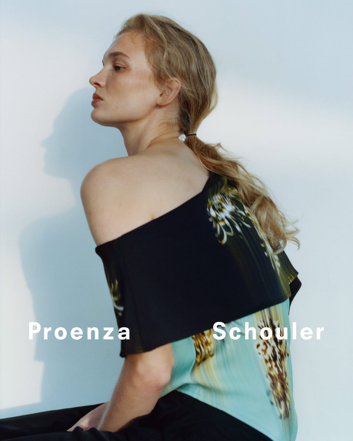 Fashion shoot of a blonde woman in a designer top and dark pants, seated against a bright white wall with the brand name displayed.