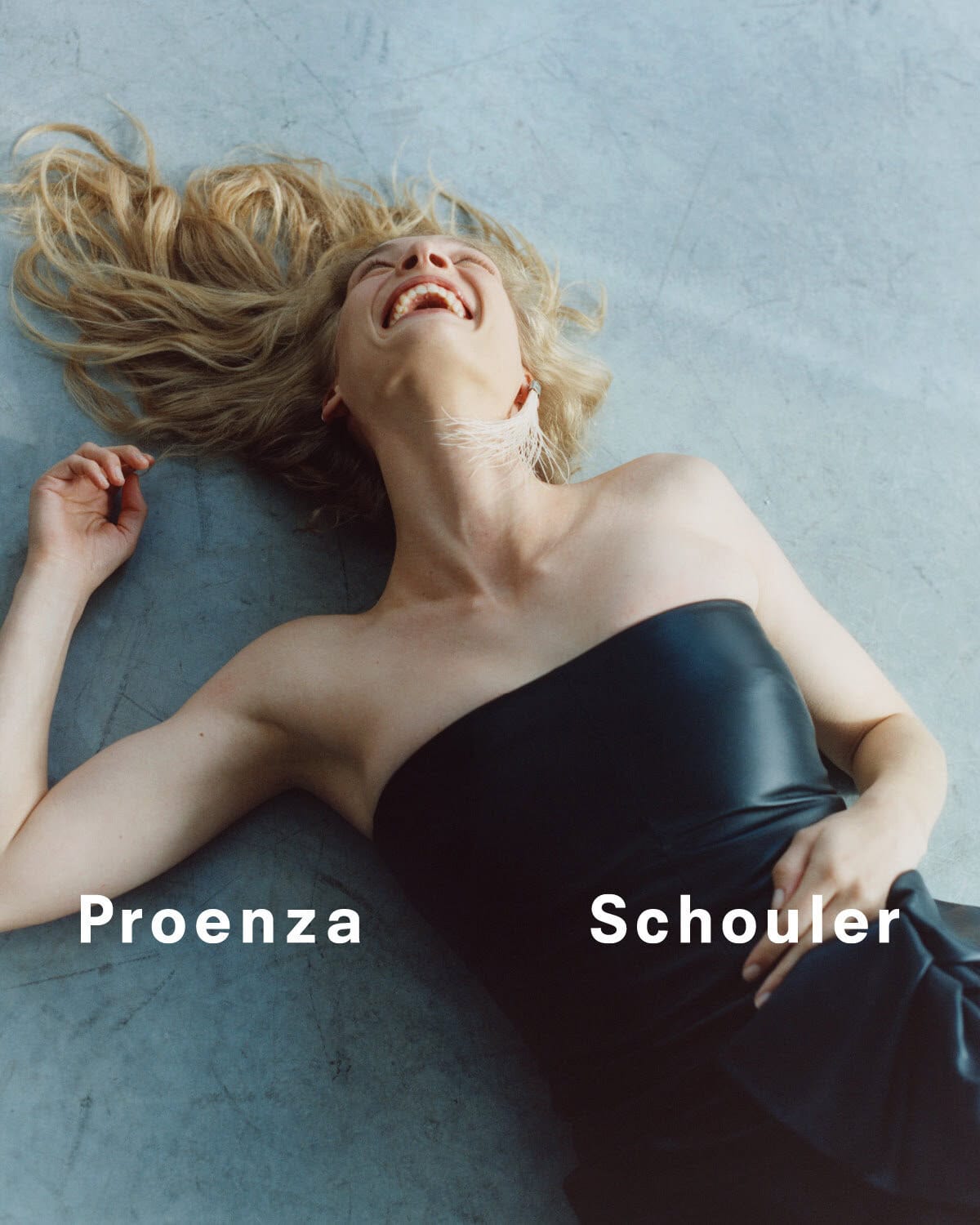 A blonde woman wearing a black dress smiles while lying on a grey surface, with Proenza Schouler text overlaid on the image.