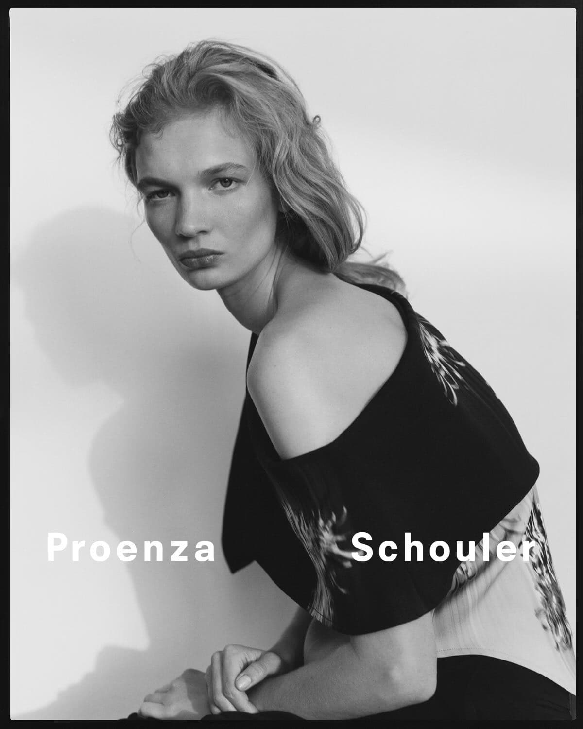 Monochrome portrait of a fair-skinned woman with blonde hair, posed in a fashion-forward black top with shoulder detail, indoors.