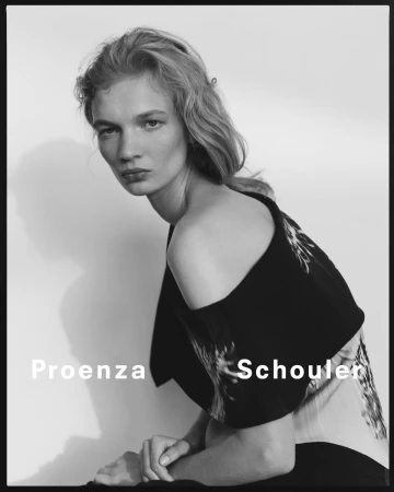 Monochrome portrait of a fair-skinned woman with blonde hair, posed in a fashion-forward black top with shoulder detail, indoors.
