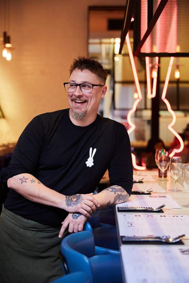 Man with glasses and tattoos smiles in a restaurant setting, leaning against a bar with menus. A neon rabbit sign glows softly.