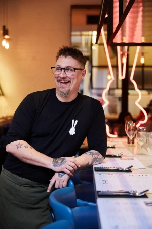 Man with glasses and tattoos smiles in a restaurant setting, leaning against a bar with menus. A neon rabbit sign glows softly.