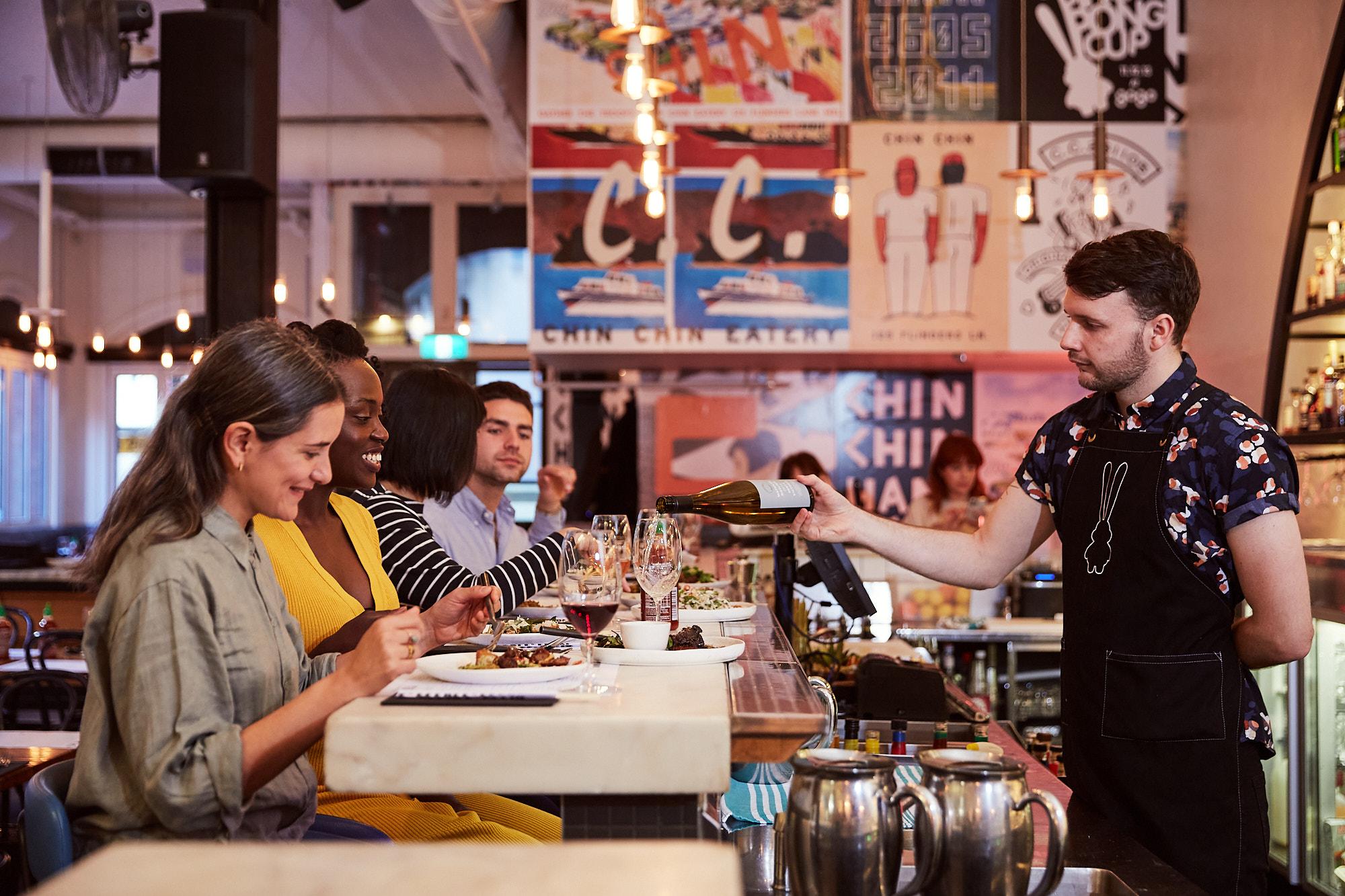 Restaurant scene featuring a bartender pouring wine for a group of patrons seated at the bar, enjoying food and drinks in a vibrant atmosphere.