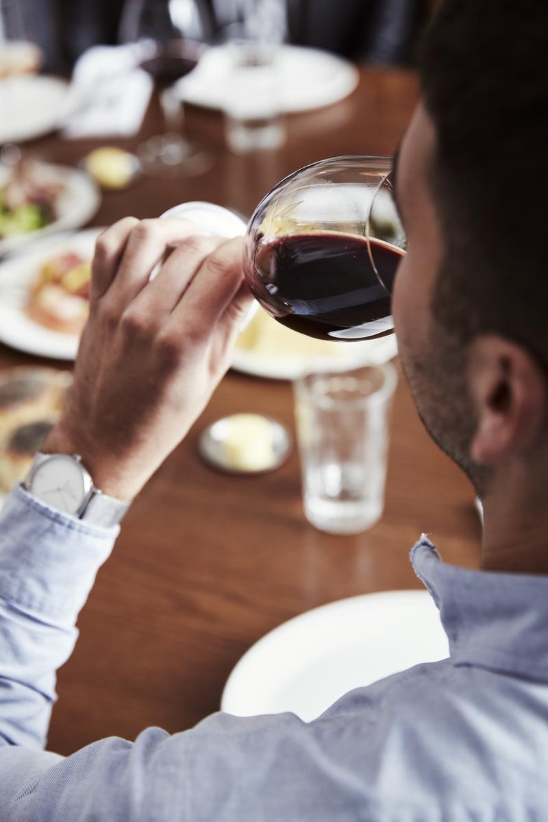 Man drinking red wine from a wine glass while seated at a table with other glasses, plates, and food, viewed from behind.