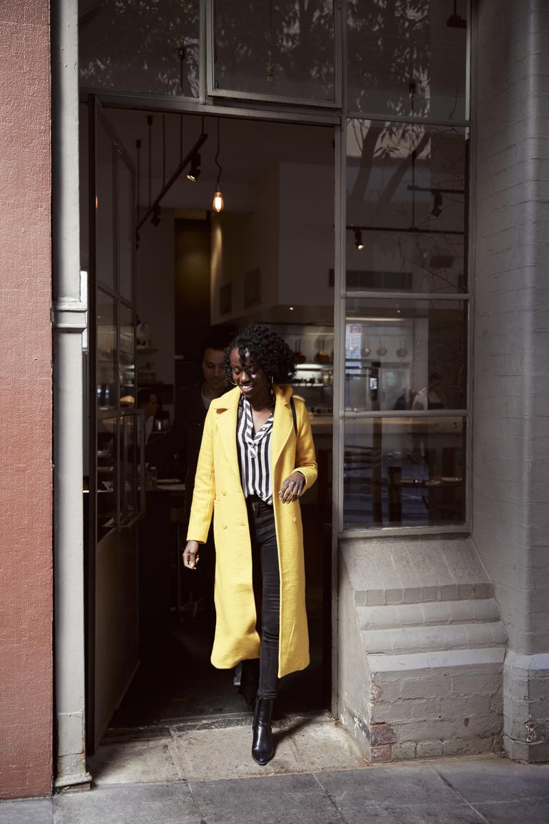 A Black woman in a yellow coat exits a building with a glass-fronted doorway, wearing black pants and boots, captured in a medium shot.