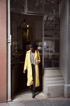 A Black woman in a yellow coat exits a building with a glass-fronted doorway, wearing black pants and boots, captured in a medium shot.