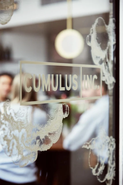 A view through a glass door with the words 'Cumulus Inc.' printed on it, with people visible inside a bright, indoor setting.