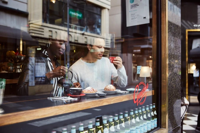 Two people at a cafe table, one pouring tea from a black teapot, the other drinking coffee; baked goods and bottled beverages visible.