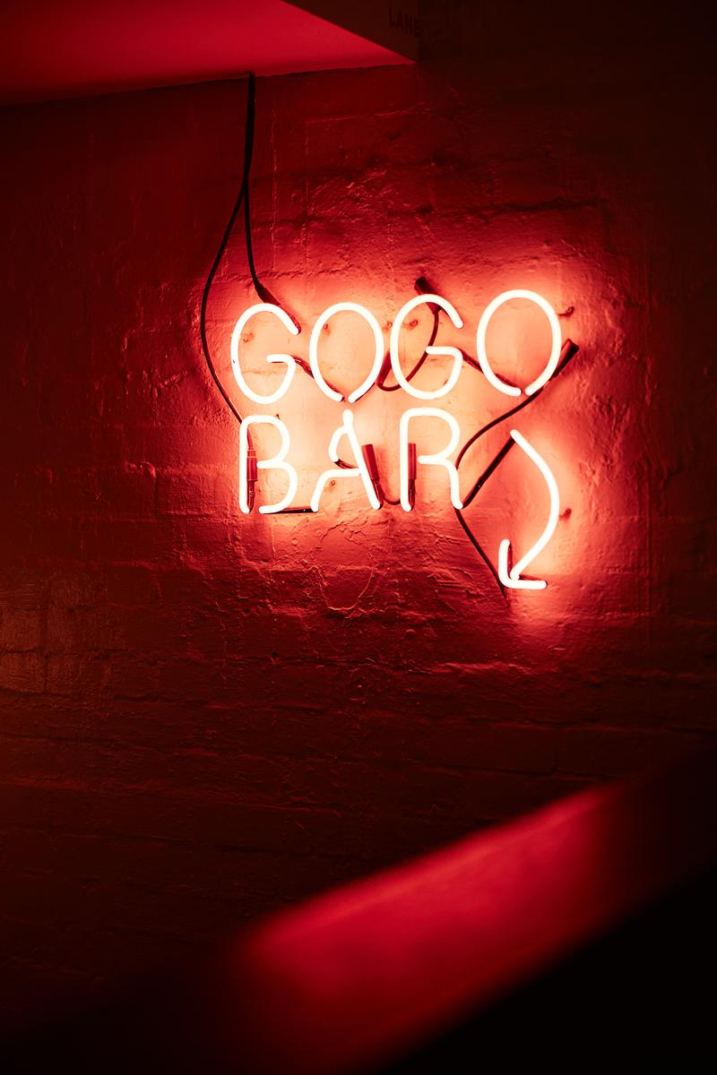 A neon sign that reads "GOGO BAR" with a downward arrow, illuminated in a dark room with a brick wall.