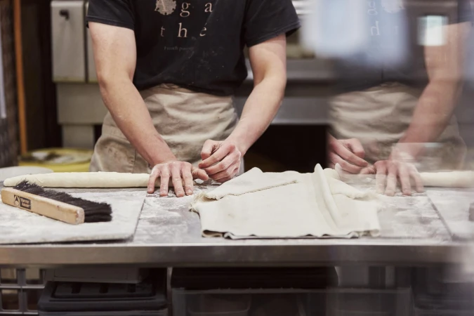 Two bakers in aprons work at a metal counter in a kitchen, shaping dough for pastries, with visible brush and flour dusting.