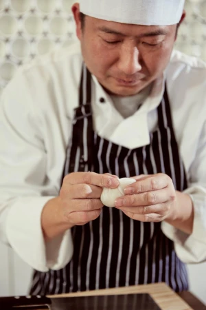 An Asian chef in uniform meticulously shapes dough, displaying expertise in a kitchen with soft lighting and a focus on culinary precision.