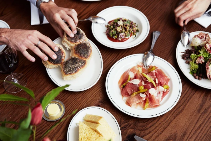 Overhead shot of a table with several plates of food, including bread, salad, and ham, along with people's hands and a glass of wine.