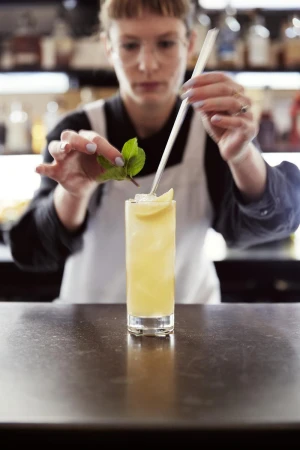 Adult female bartender prepares a yellow cocktail with ice, mint leaves, and a straw, at a restaurant counter with blurred shelves.