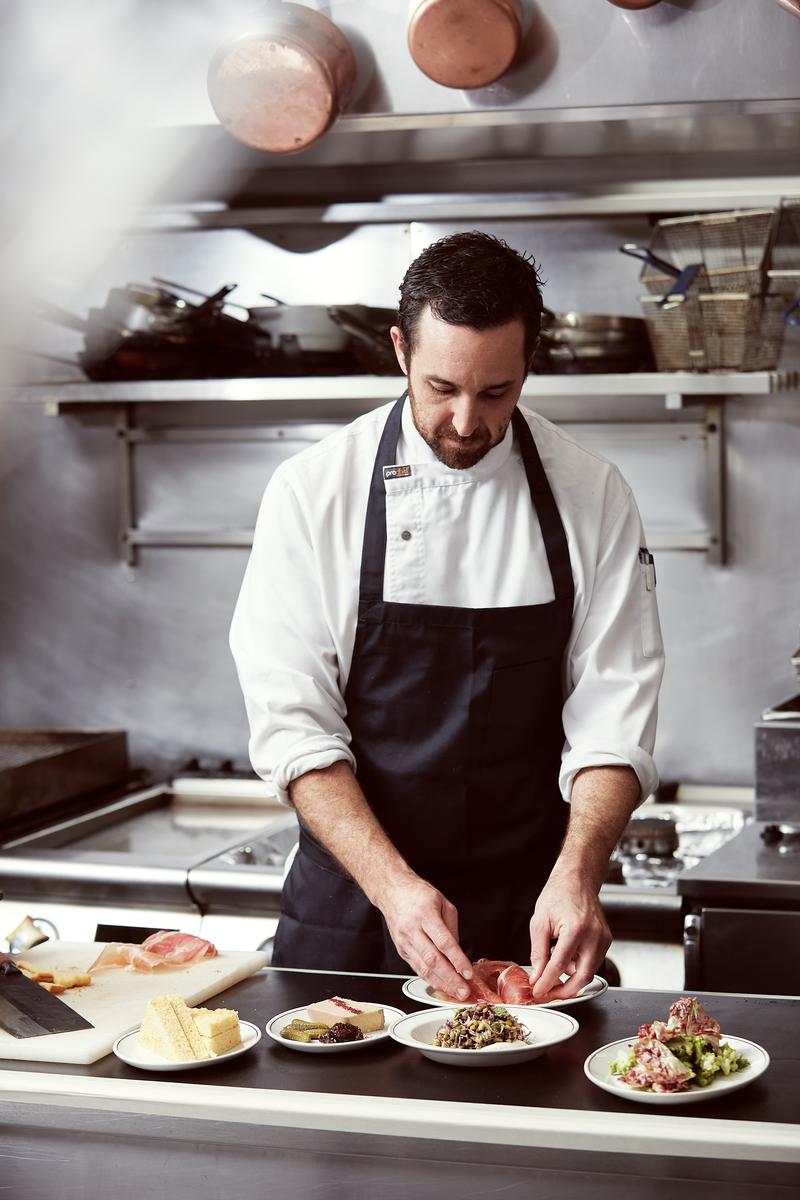 Male chef in a white uniform and black apron prepares small plates of food on a stainless-steel kitchen counter with metal pans above.