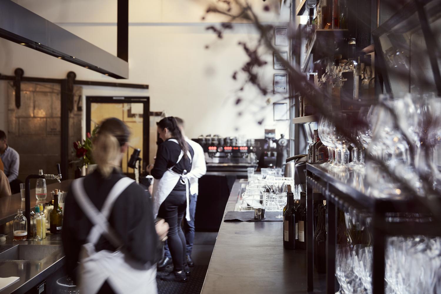 Busy restaurant interior with bartenders, patrons at the bar, visible shelves of glassware, and dim lighting.