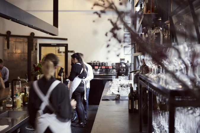 Busy restaurant interior with bartenders, patrons at the bar, visible shelves of glassware, and dim lighting.