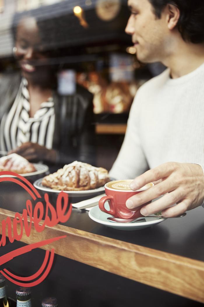 Man at cafe with coffee and croissant, an African-descent woman visible in the background, indoor setting, natural light.