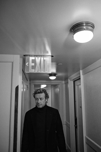 Monochrome portrait of a man in a dark coat and turtleneck standing in a hallway with an exit sign overhead.